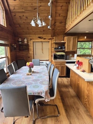 Interno di una baita in legno con tavolo da pranzo, sedie, angolo cottura e lampadario.