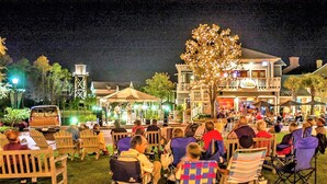 An outdoor gathering with people seated on lawn chairs, a stage with performers, and a building with a balcony in the background. - Enjoy your very own private courtyard! FREE TRAM ! Mins. to pool,dining,shopping (MIRAMAR BEACH)