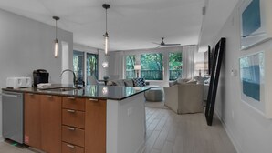 A modern kitchen with a central island, a dining area with a sofa, and a large window offering a view of trees. - Enjoy your very own private courtyard! FREE TRAM ! Mins. to pool,dining,shopping (MIRAMAR BEACH)