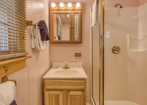 A bathroom with a wooden vanity, a mirror with lights, a glass shower enclosure, and a window with blinds.