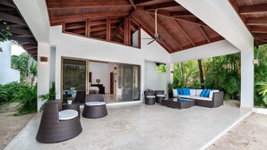 Un espacio exterior techado con mobiliario de mimbre y un sofá blanco con cojines azules.