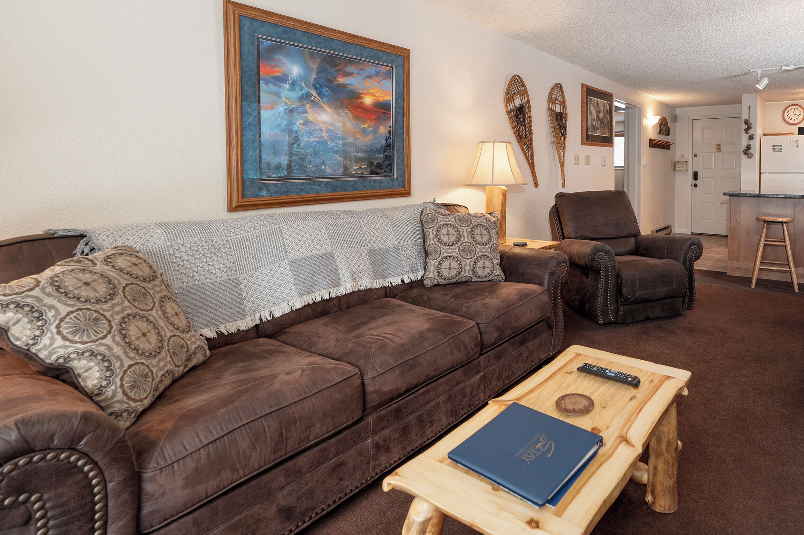 A living room with a brown leather sofa, patterned cushions, a wooden coffee table with a book, a remote control, and a painting on the wall.