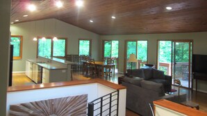 Un amplio espacio de sala con techo alto, vigas de madera, un comedor con sillas y mesa, y ventanas grandes que ofrecen vistas a follaje.