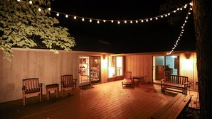 Una terraza de madera con luces colgantes, bancos y una puerta de vidrio que da a un espacio interior.