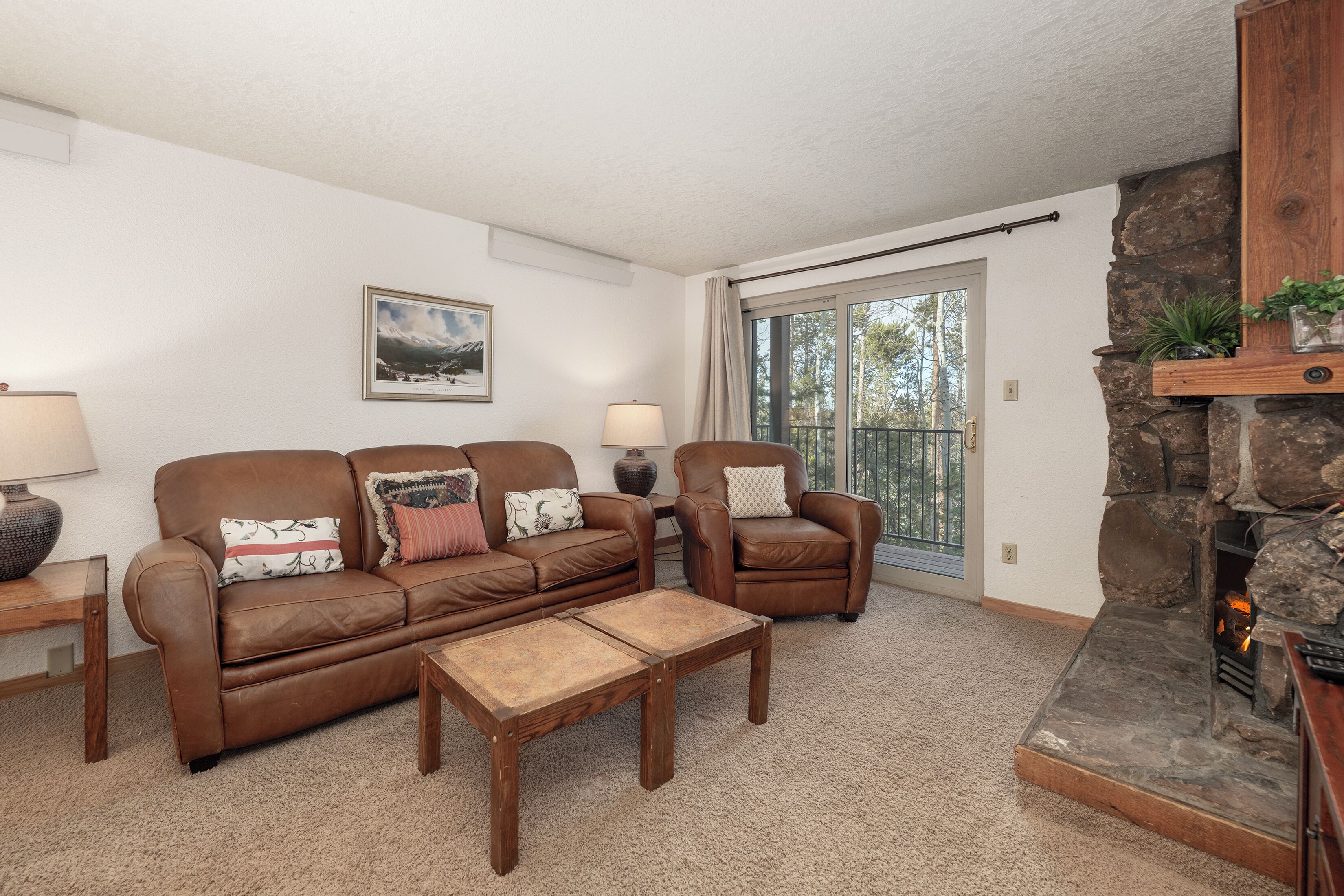 A cozy living room with a brown leather sofa, two armchairs, a coffee table, a fireplace, and a large window with a view of trees.