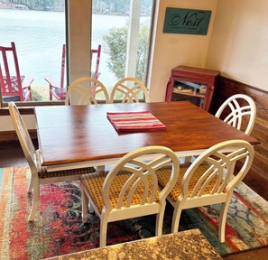 A dining area with a wooden table and white chairs, a rug, and a view of a lake through a window. - Beautiful East Texas Cottage on Private Lake - VRBO Premier Property (Holly Lake Ranch)