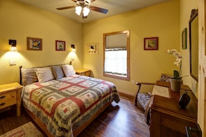 A bedroom with a bed, nightstands, a desk, a chair, a window with blinds, a ceiling fan, and wall-mounted lights.