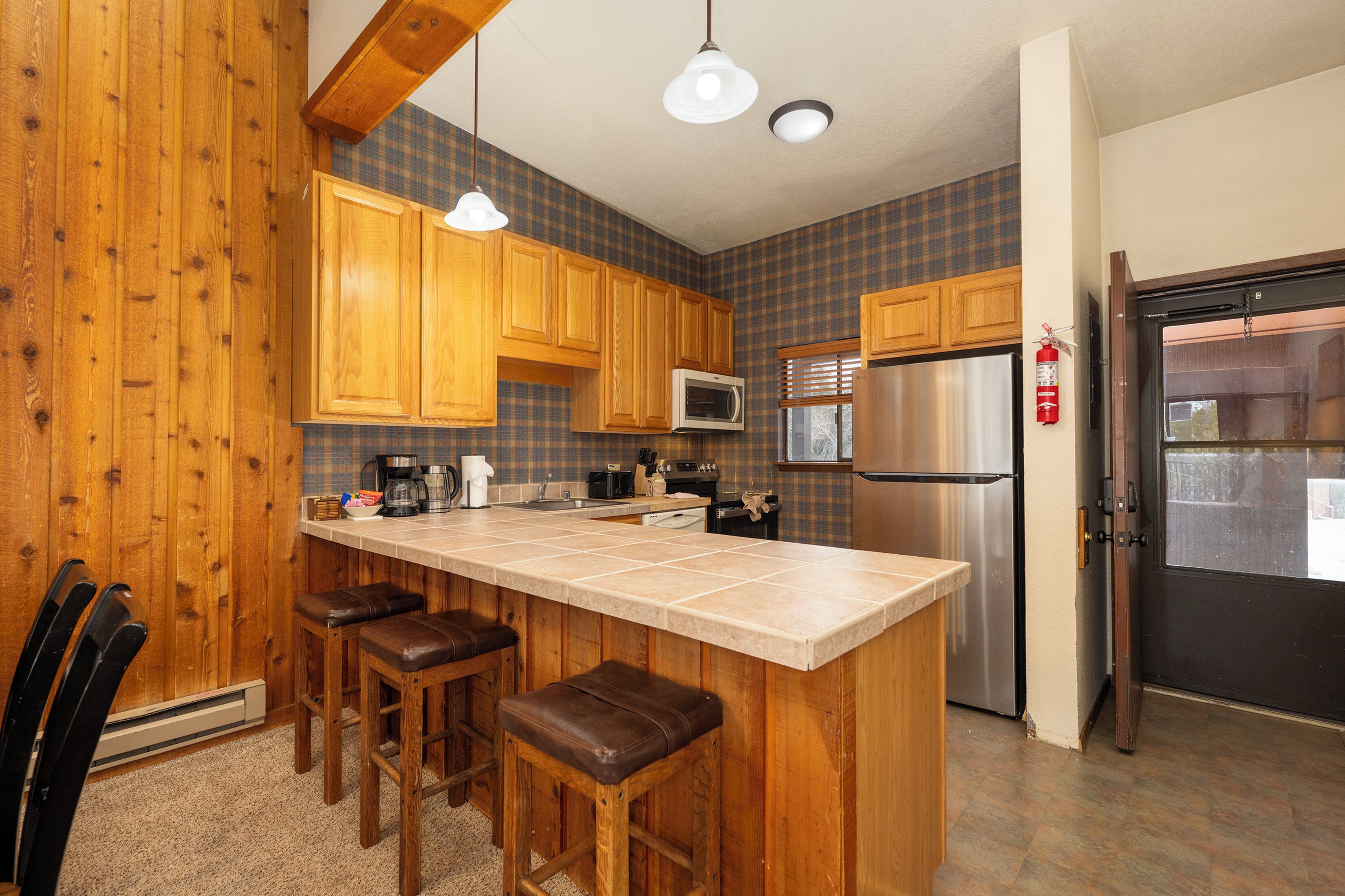 A kitchen with wooden cabinets, a stainless steel refrigerator, a microwave, and a bar stool area.