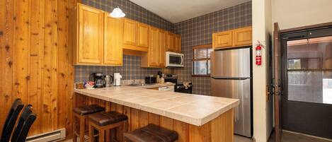 A kitchen with wooden cabinets, a stainless steel refrigerator, a microwave, and a bar stool area.