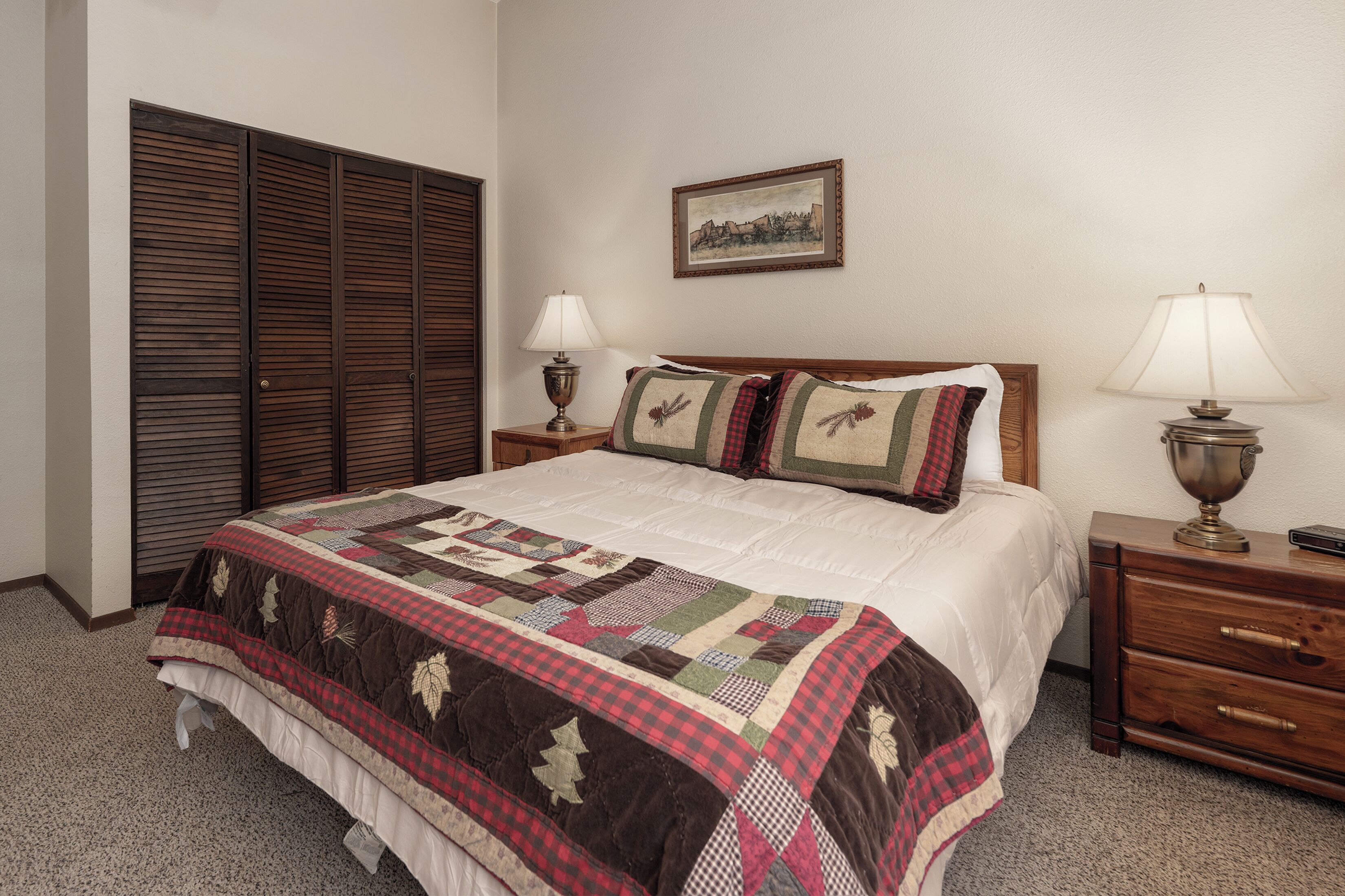 A neatly made bed with a quilt, two bedside tables with lamps, a wooden nightstand, and a large closet with sliding doors.