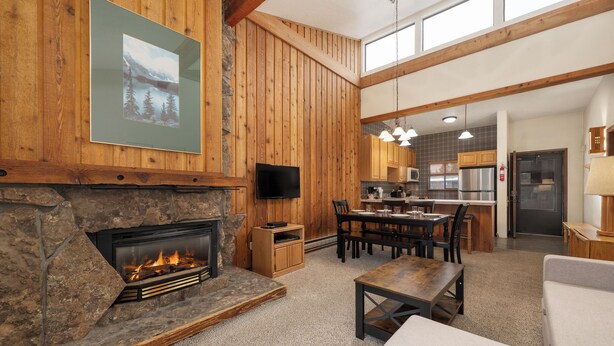 A spacious living area with a stone fireplace, wooden walls, a dining table, and a kitchen in the background.