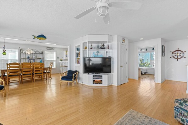 A spacious living room with a flat-screen TV, wooden flooring, and a dining area with chairs and a table. - ☀️Casablanca: Oceanfront Home w/ Ocean Views☀️ (Kure Beach)