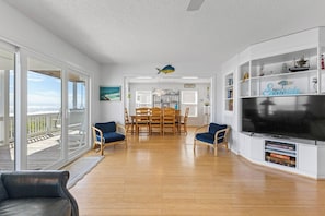 A spacious living room with a large TV, wooden flooring, and a dining area with chairs and a table. - ☀️Casablanca: Oceanfront Home w/ Ocean Views☀️ (Kure Beach)