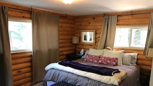 Ein Schlafzimmer in einer Blockhütte mit einem Bett, Nachttischen, einer Bank und einem Fenster mit Vorhängen.