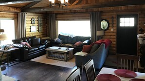Ein gemütliches Blockhaus-Interieur mit Holzbalkendecke, einem Ledersofa, einem Esstisch mit Platzsets und einem großen Fenster, das natürliches Licht hereinlässt.