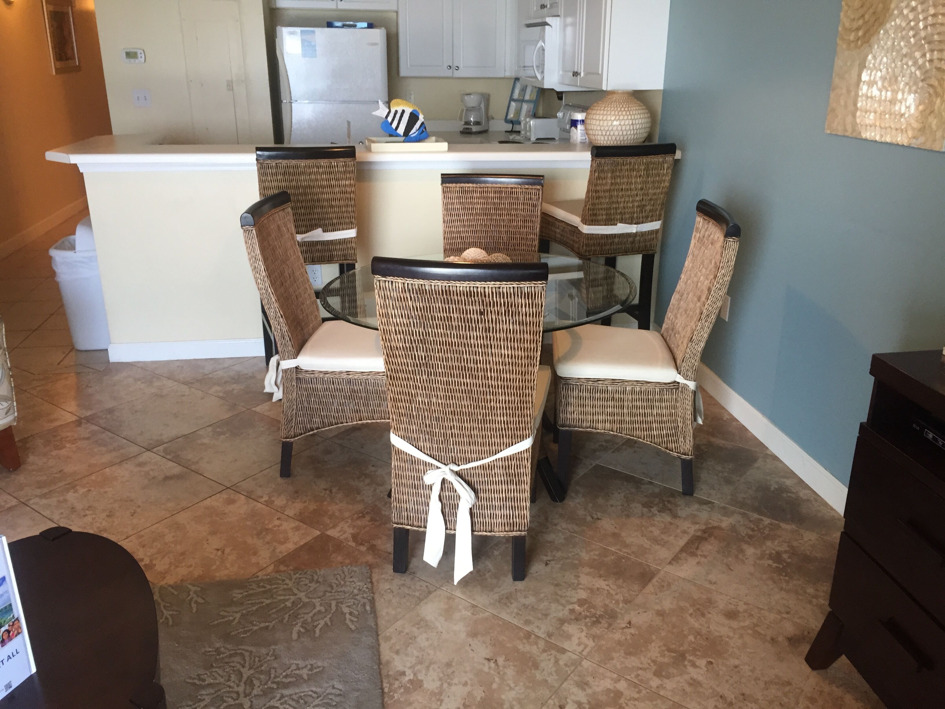 Un coin repas avec des chaises en osier et une table en verre, une cuisine à l’arrière-plan et un sol carrelé.