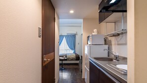 Een smalle gang die leidt naar een slaapkamer met een bed, een klein tafeltje en een raam met gordijnen. Een kitchenette met een magnetron, spoelbak en kastjes is aan de rechterkant zichtbaar.