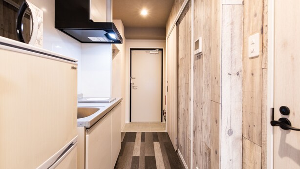Un pasillo estrecho con una zona de cocina a un lado, que incluye un refrigerador, una estufa y un lavamanos. El pasillo tiene piso de madera y una puerta al final.