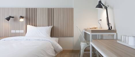 A modern hotel room with a wooden headboard, a bed with white linens, a desk with a lamp, and a chair.