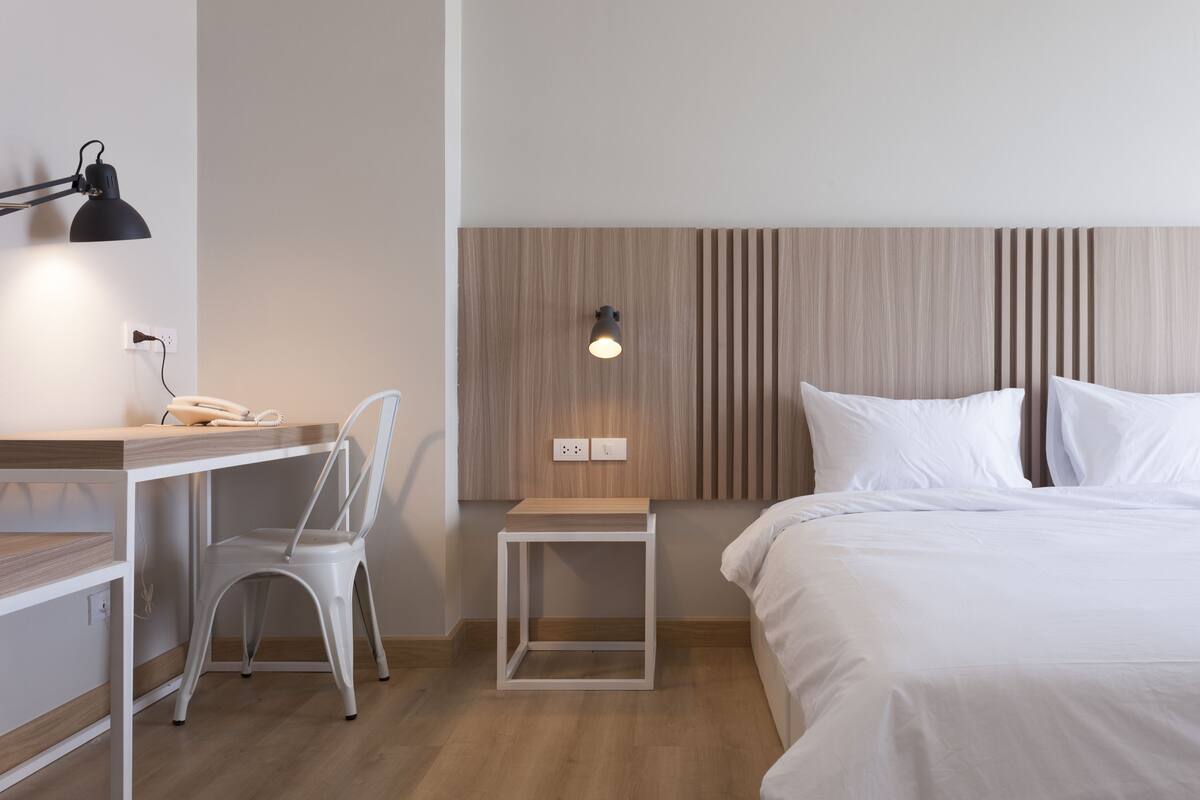 A hotel room with a bed, a desk with a chair, a small table, a lamp, and a wall-mounted light.