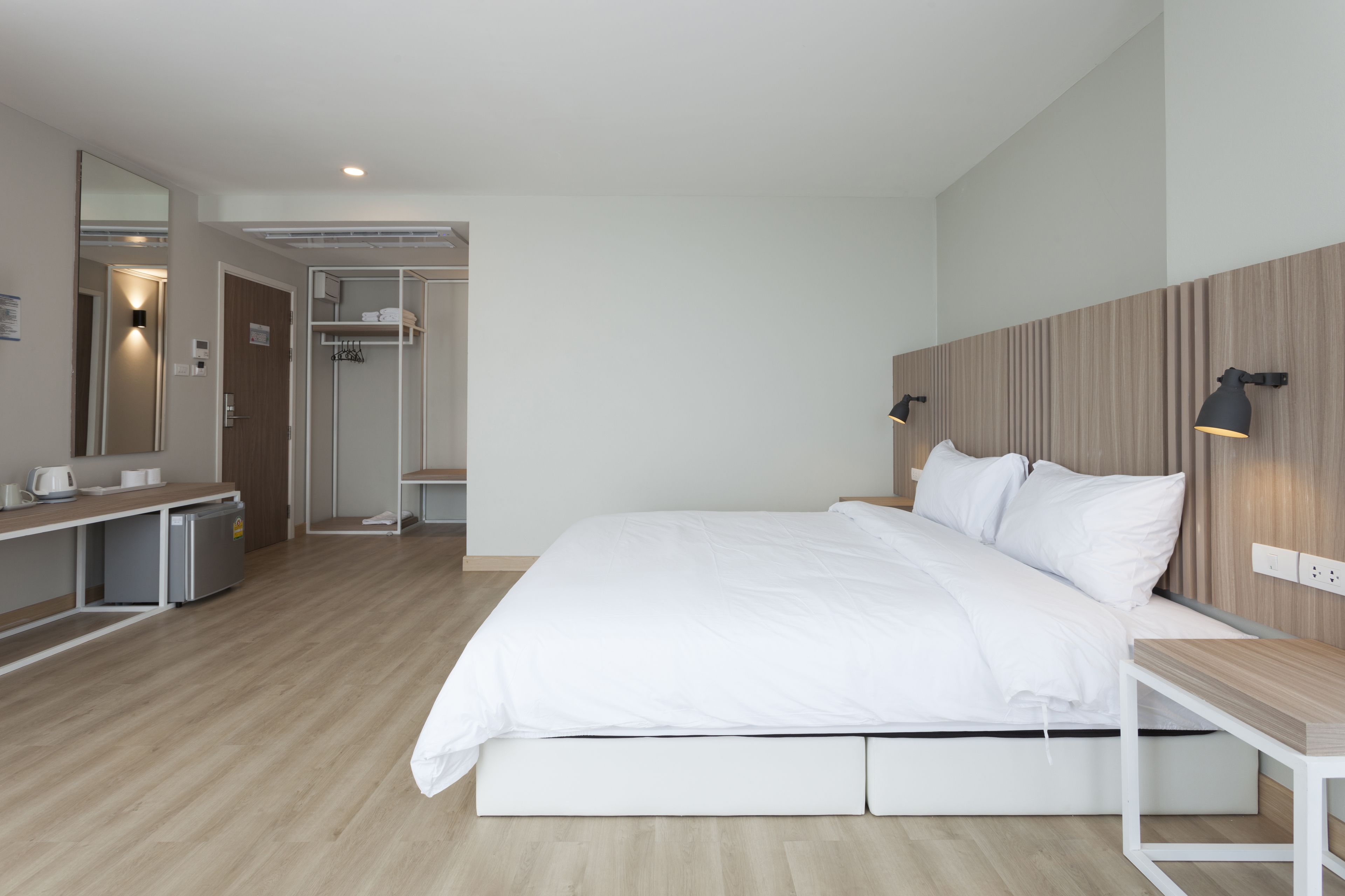 A modern hotel room with a bed, bedside table, and a mirror. There is a small desk with a chair and a refrigerator. The room has wooden flooring and a wooden panel wall.