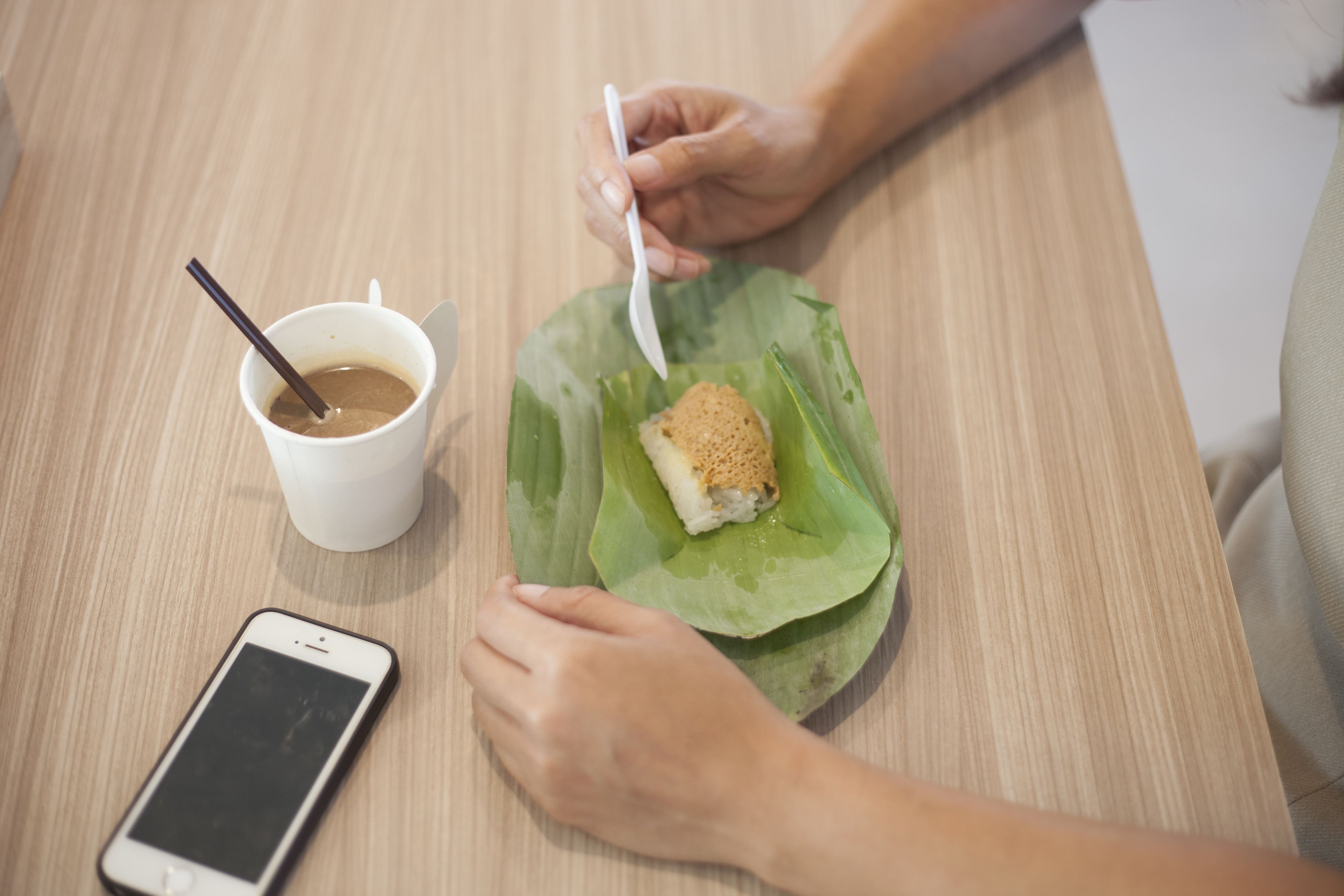 A person eating a sandwich on a banana leaf with a cup of coffee and a spoon on a wooden table.