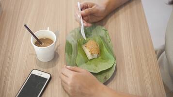 A person eating a sandwich on a banana leaf with a cup of coffee and a spoon on a wooden table.