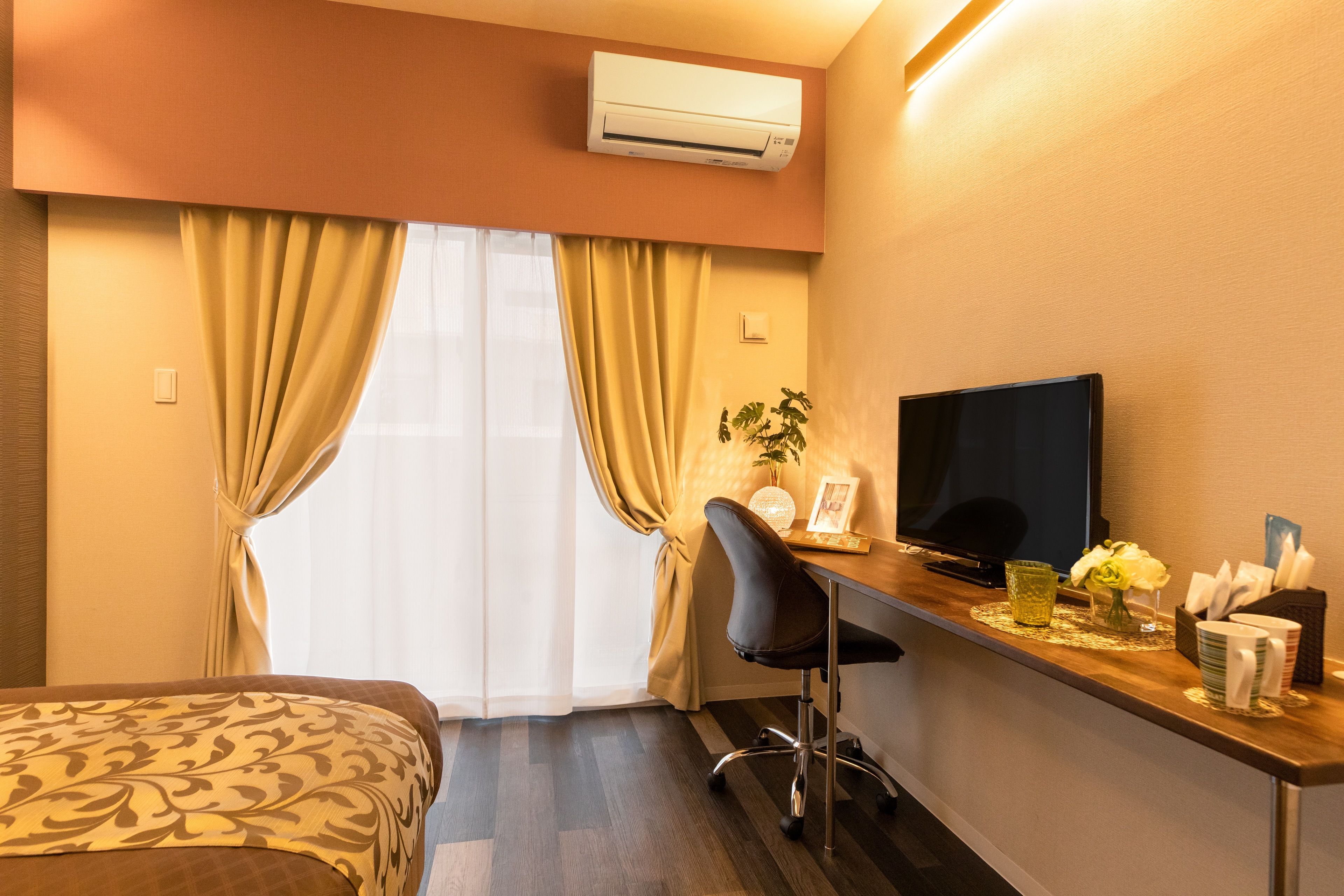 A hotel room with a bed, a desk with a television, a chair, and a small table with a vase and a glass.