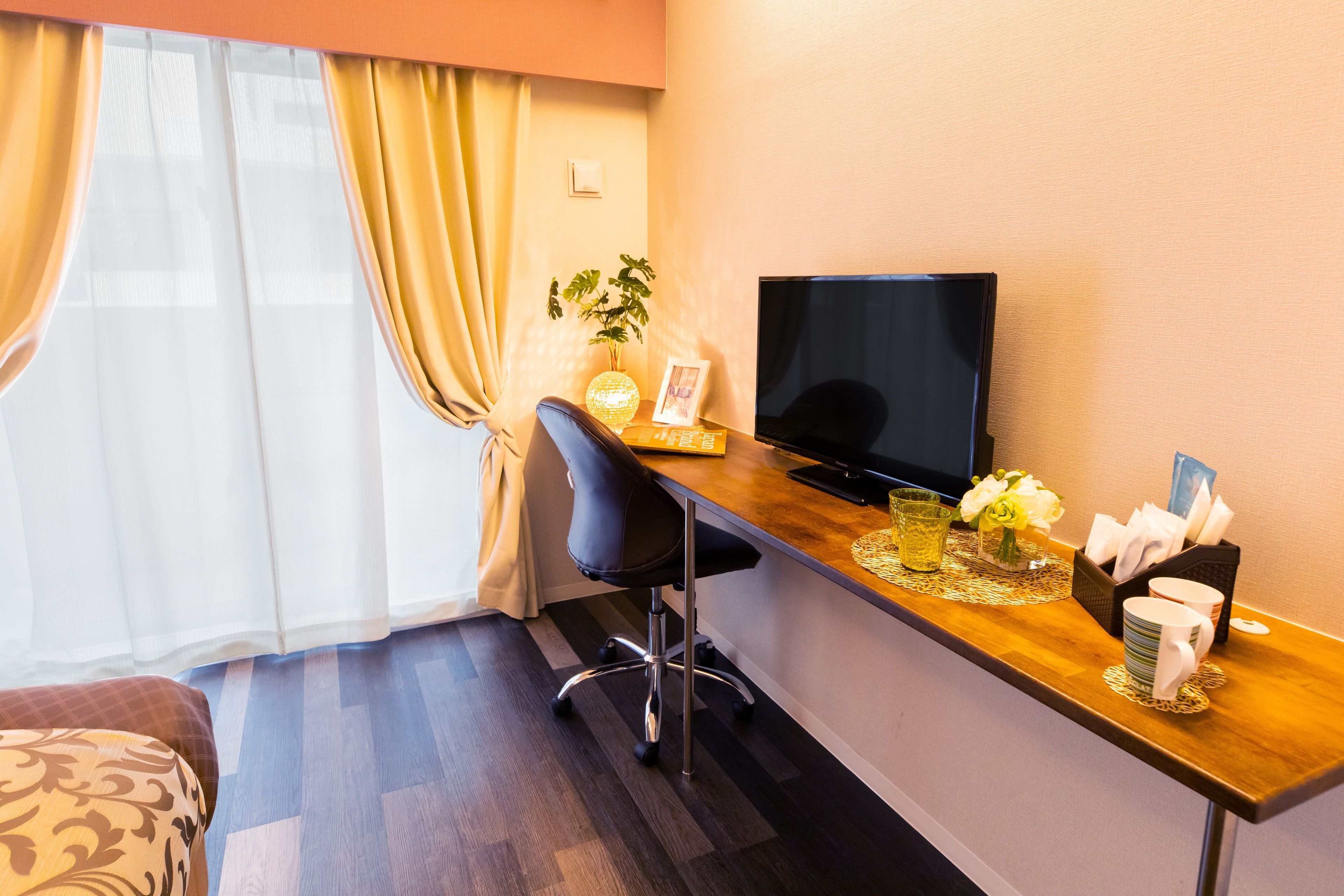A hotel room with a wooden desk, a black chair, a television, a potted plant, and a window with curtains.