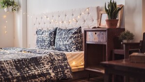 Ein Schlafzimmer mit einer gepolsterten Kopfteile, gemusterten Betttextilien, Lichterketten, einem Nachttisch aus Holz und einer Topfpflanze.