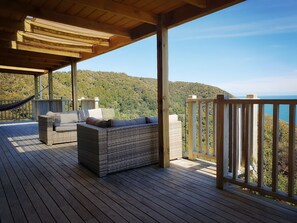 Una terraza de madera con un sofá de mimbre y una hamaca, con vistas a un paisaje montañoso.