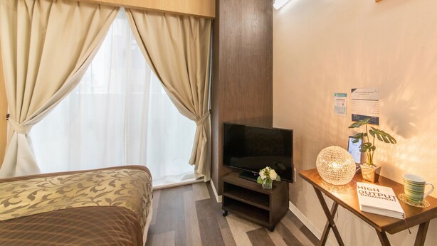 A hotel room with a bed, a TV, a desk with a book, and a plant. - Mr.KINJO in UCHIDOMARI (Ginowan)