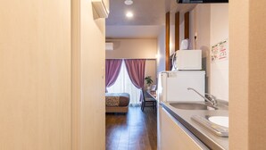 A compact hotel room with a kitchenette, a bed with a patterned headboard, and a window with purple curtains. - Mr.KINJO in ISHIKAWA inter (Uruma)