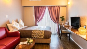 A hotel room with a bed, a red sofa, a wooden table with a vase of flowers, a television, and a desk with a lamp. - Mr.KINJO in ISHIKAWA inter (Uruma)