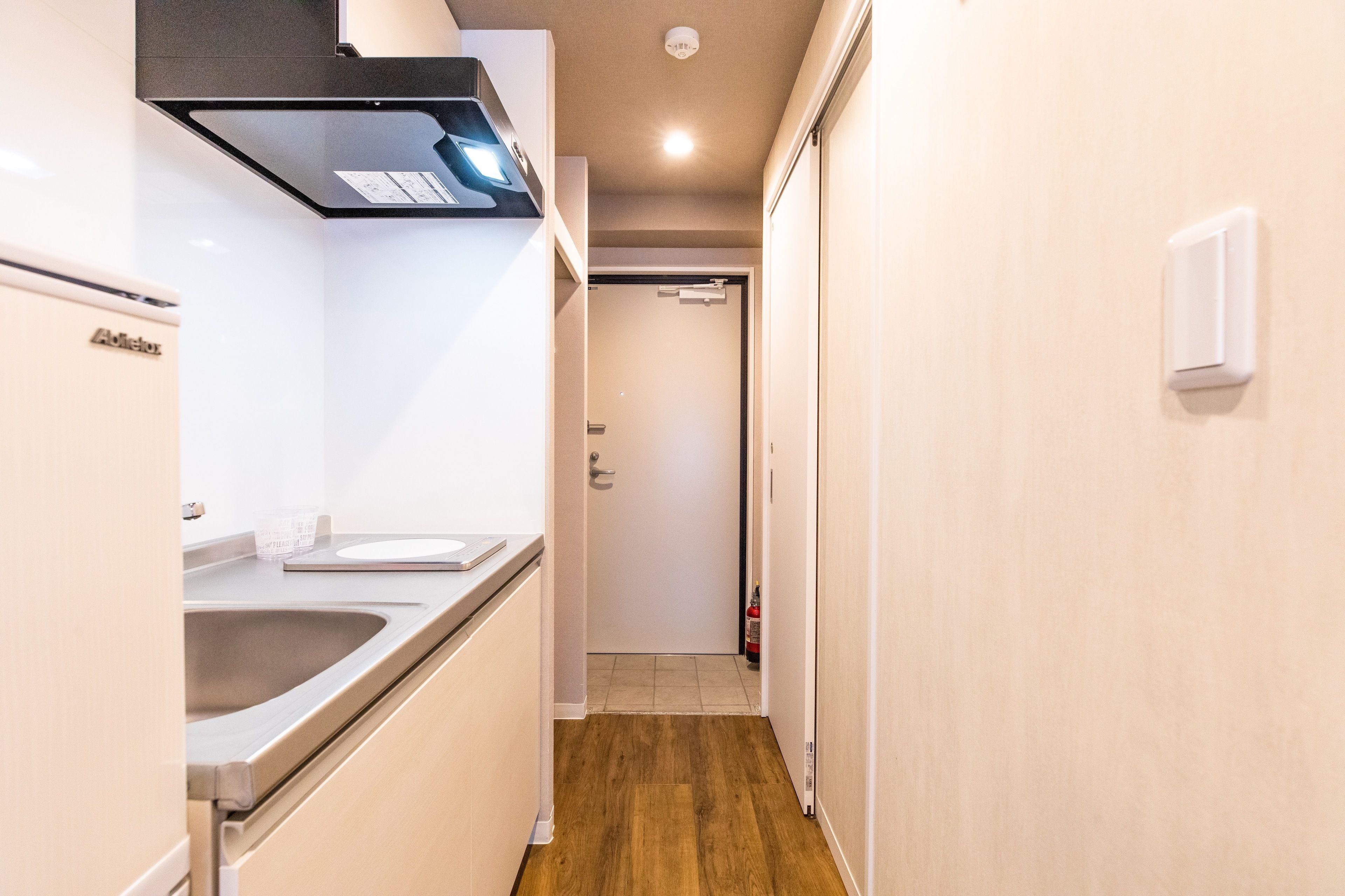 Un pasillo estrecho con una zona de cocina a la izquierda, que incluye un lavamanos de acero inoxidable y un refrigerador blanco. Un extintor está montado en la pared cerca de la puerta.