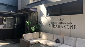Un rincón con asientos, un sofá y una mesa de vidrio, un cartel que indica el Hotel Yumoto Station MIRAHAKONE y la entrada del edificio con una puerta y una ventana.