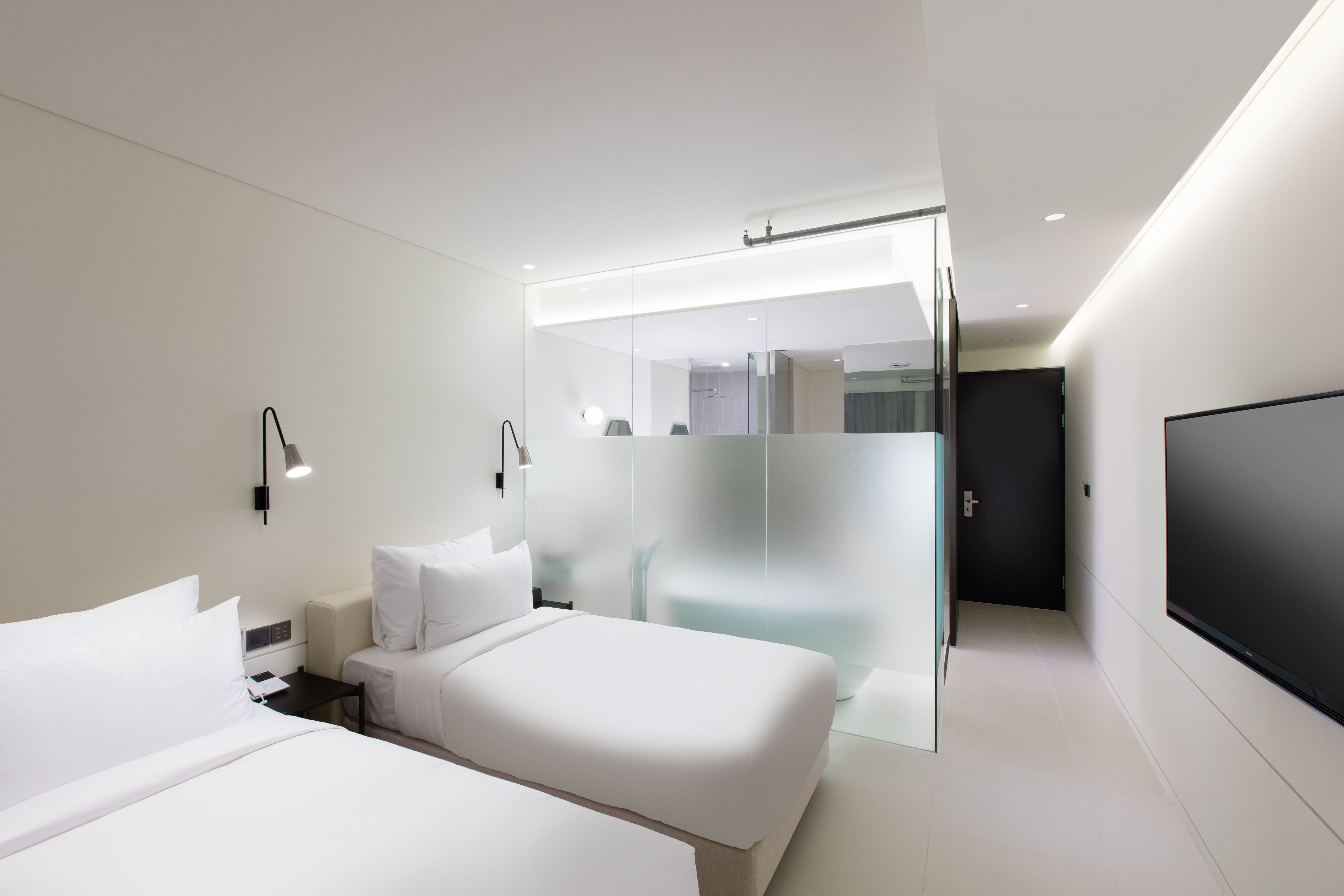Una habitación de hotel moderna con una cama grande, mesitas de noche, un televisor y un muro divisorio de vidrio.