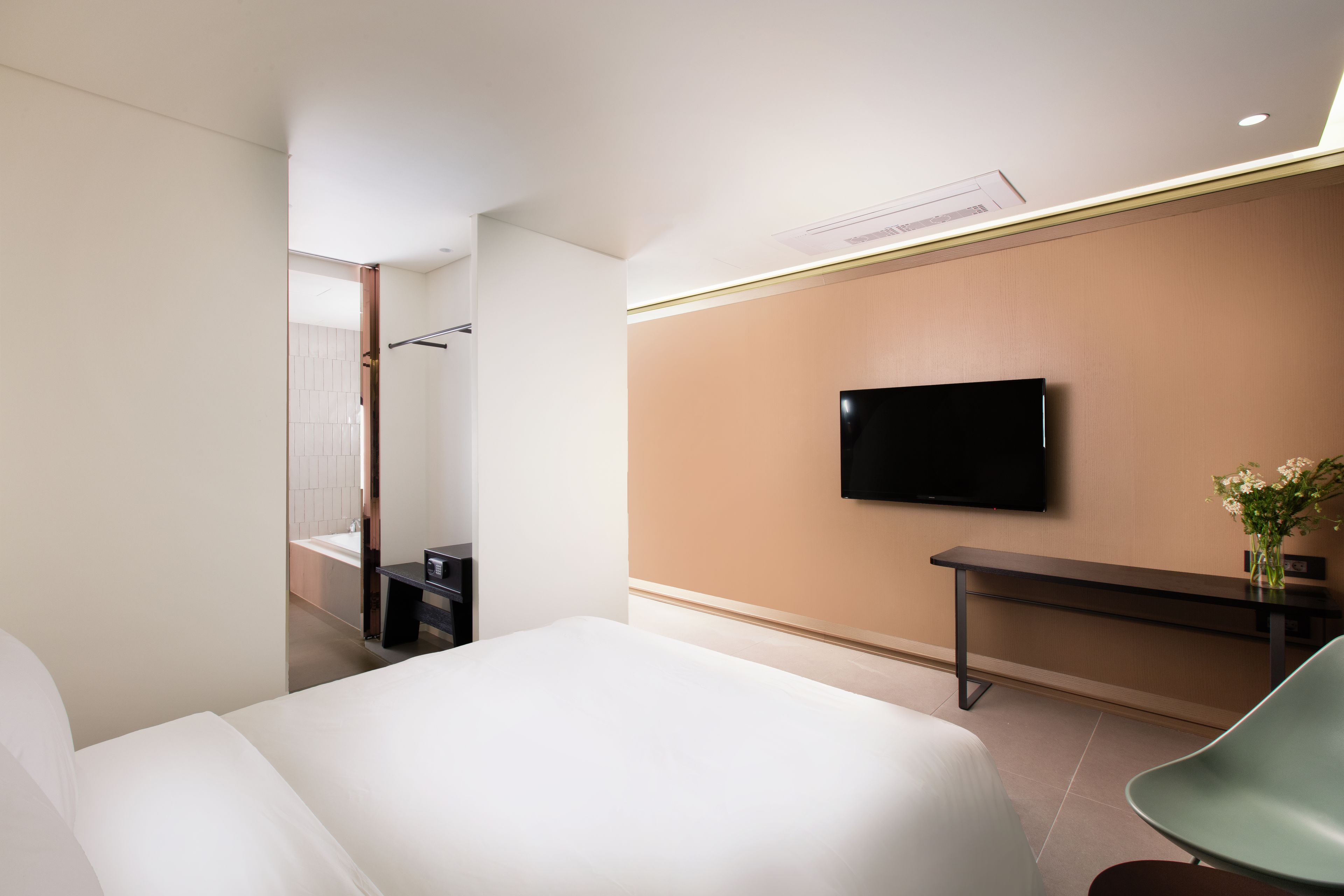 A modern hotel room with a large bed, a flat-screen TV mounted on the wall, a small desk, and a green chair.