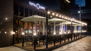 Un café al aire libre con mesas y sombrillas, fachada de vidrio y un letrero que dice 'Total Caffe'.