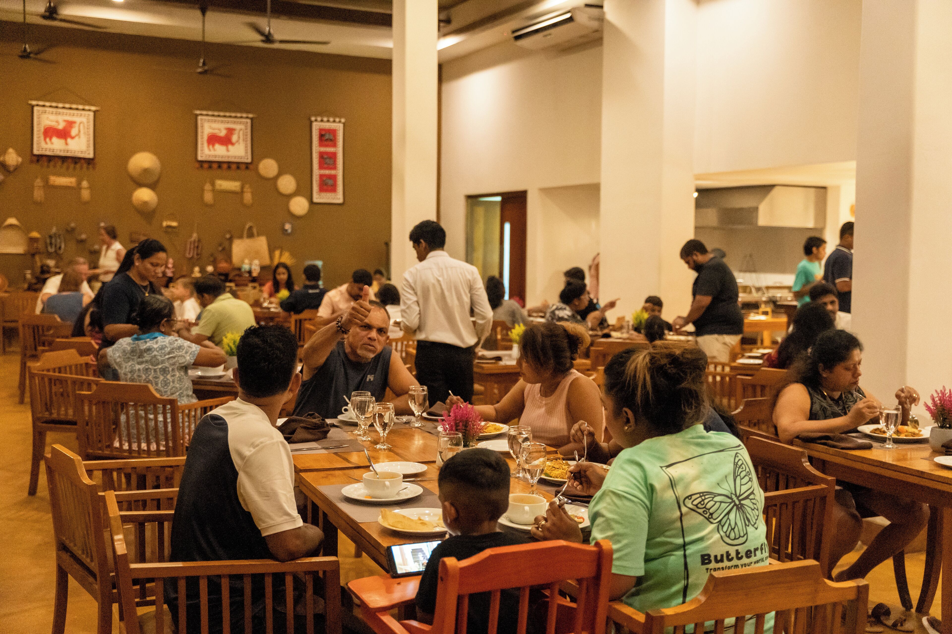A busy restaurant with wooden tables and chairs, patrons dining, and a kitchen area in the background.