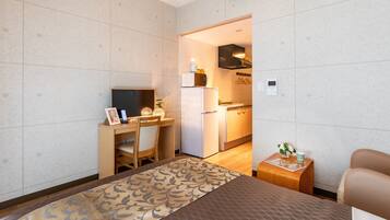 A hotel room with a bed, a desk, a chair, a small table with a vase, and a kitchenette in the background.