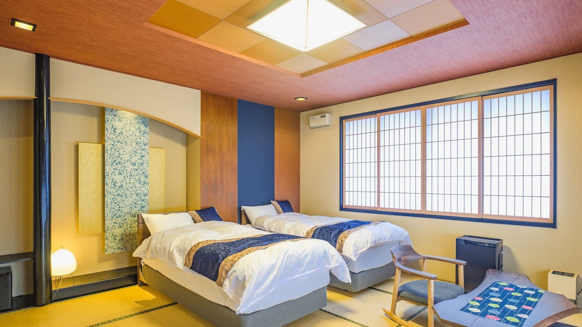 Une chambre d’hôtel avec deux lits, une grande fenêtre avec des stores à lamelles, un bureau avec une chaise et un sol recouvert de tatamis, des nattes traditionnelles japonaises.