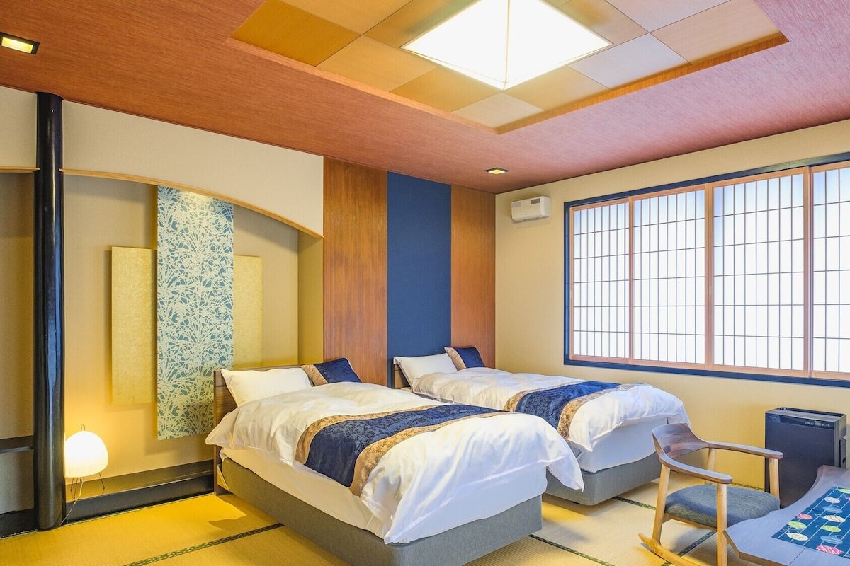 A hotel room with two beds, a large window with grid blinds, a desk with a chair, and a traditional Japanese tatami mat floor.