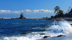 A coastal scene with waves crashing onto the shore, a rocky beach, and a mountain range in the background. - Asahiya (Himi)