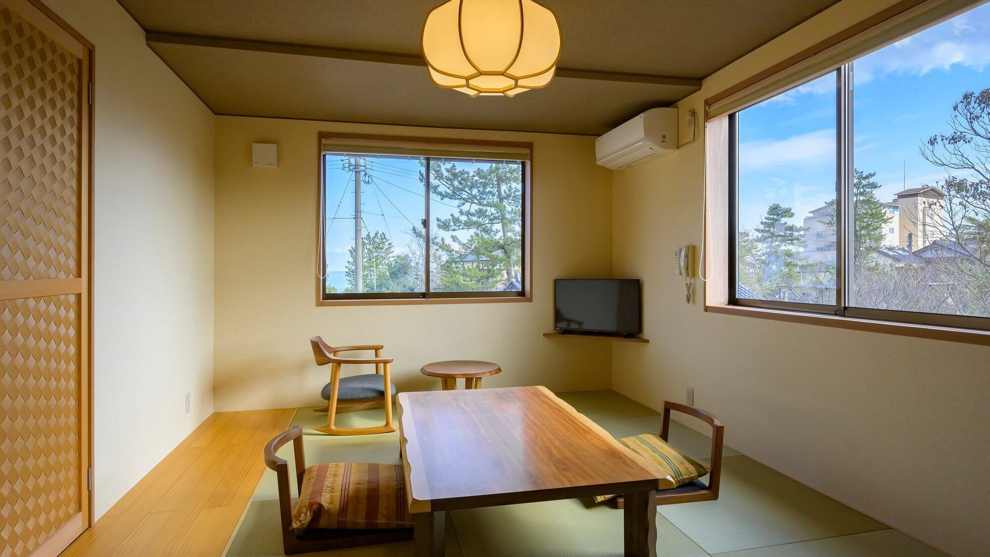 Une pièce avec un sol recouvert de tatamis, une table en bois, un téléviseur et une fenêtre donnant sur des arbres et des immeubles.