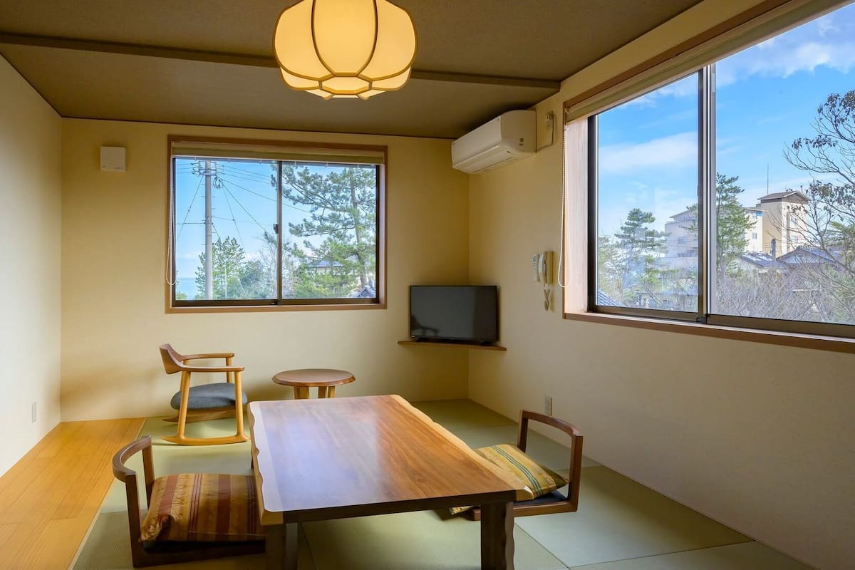 A room with tatami flooring, a wooden table, a television, and a window with a view of trees and buildings.