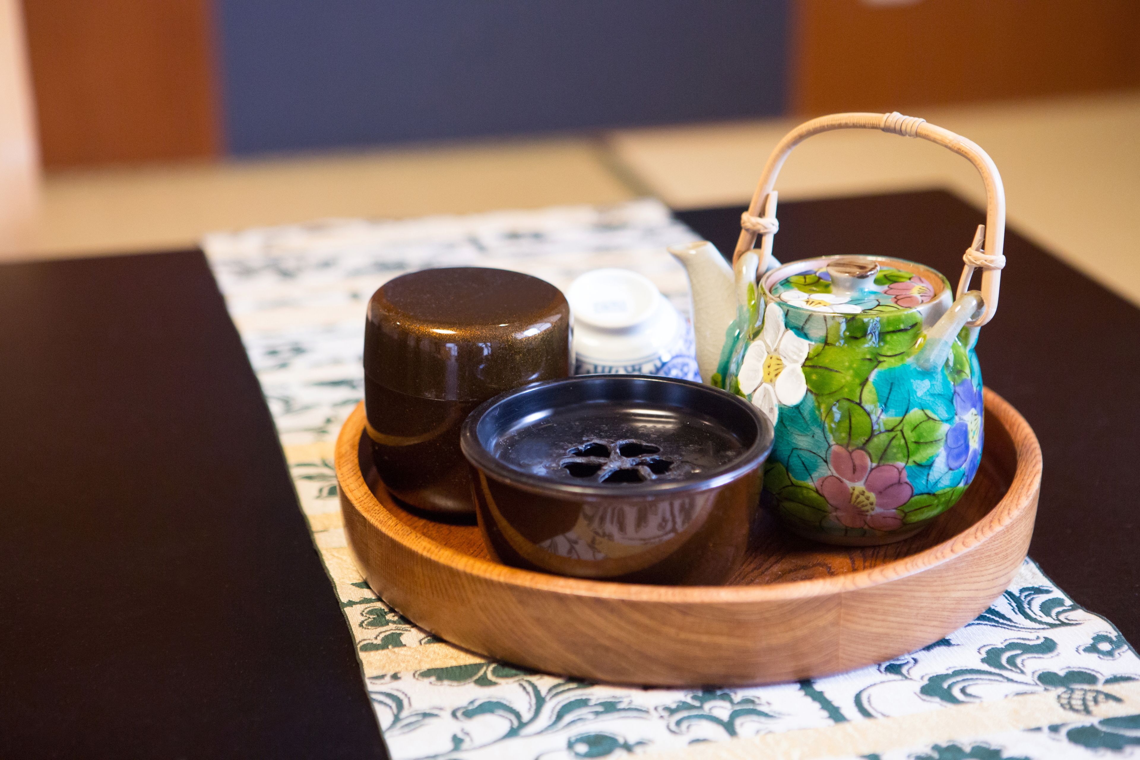 A wooden tray with a decorative teapot, a small bowl, and a bottle on a patterned tablecloth.