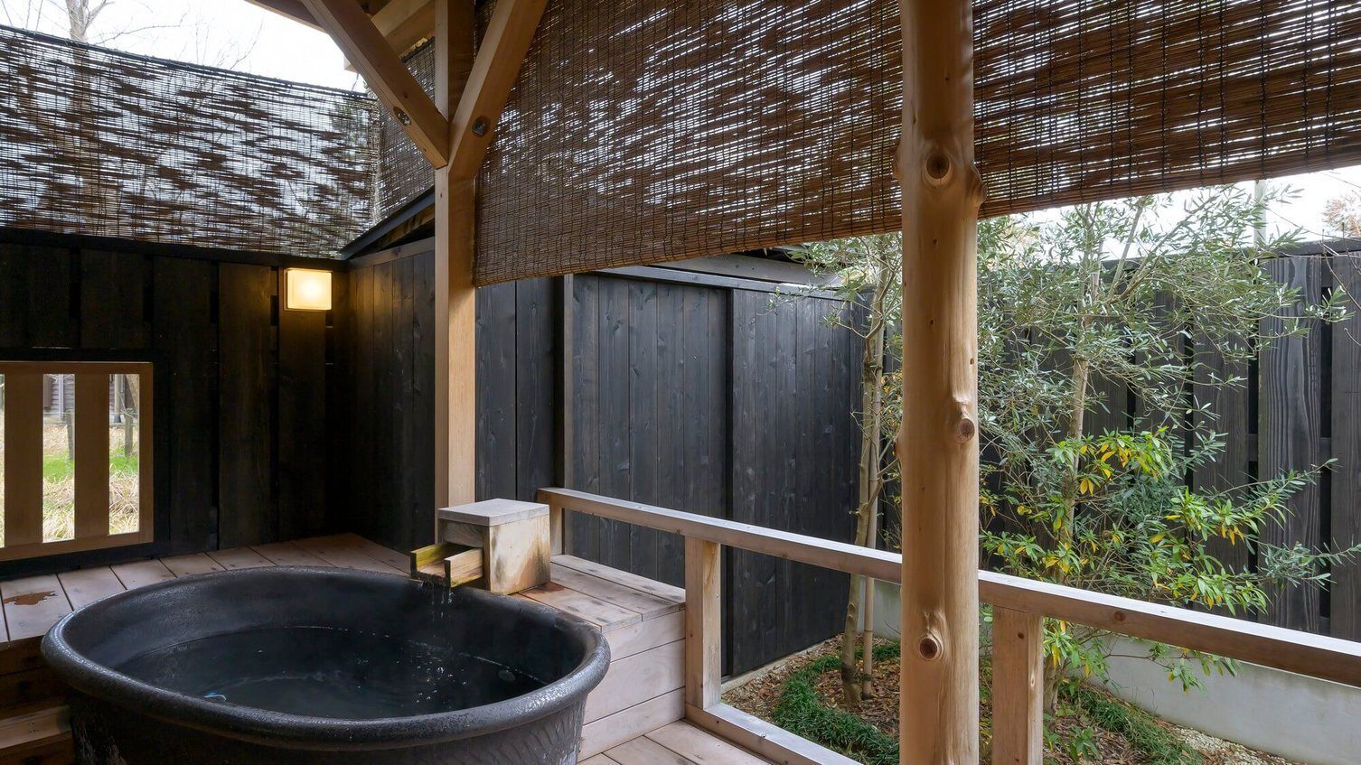 Une terrasse en bois avec un jacuzzi, des stores en bambou et une vue sur un jardin.