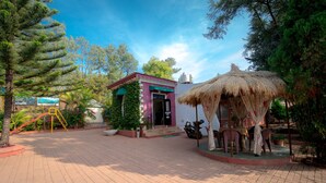 A paved courtyard with a small building, a thatched structure, and surrounding greenery. - Umbrella Inn (Mahabaleshwar)