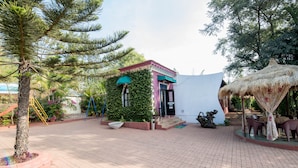 A small building with a pink roof and green foliage, surrounded by a paved area with a thatched structure and outdoor seating. - Umbrella Inn (Mahabaleshwar)